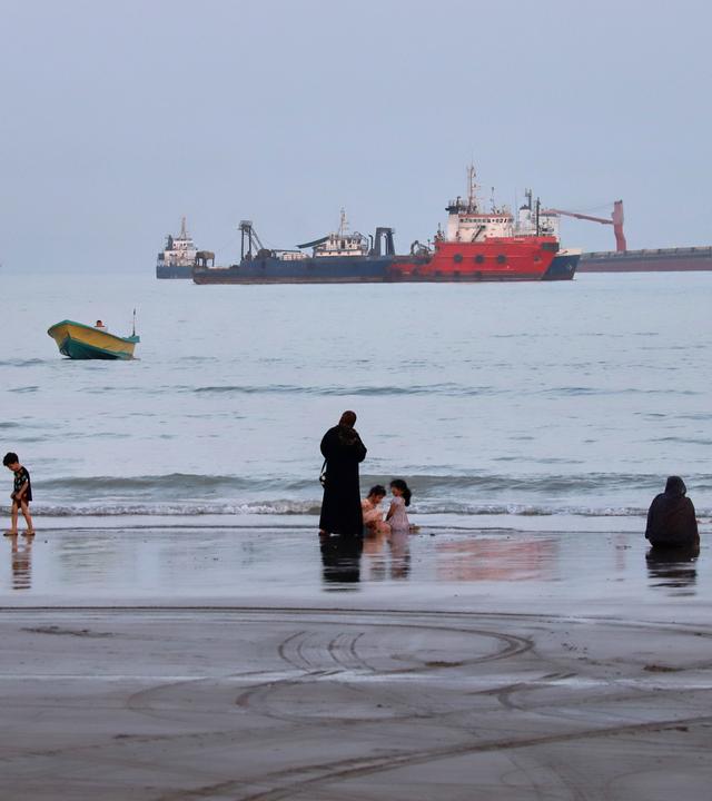 Menschen verbringen Zeit am Strand, während vor Bandar Abbas im Iran in der Straße von Hormus Handelsschiffe vor Anker liegen