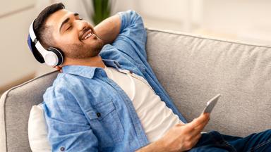 Ein Mann liegt auf einem Sofa und hört Musik über Kopfhörer und Samrtphone.