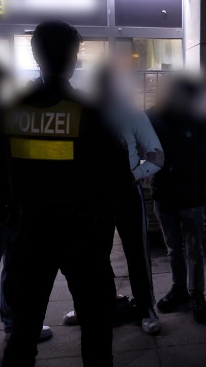 Zwei Polizeibeamten auf Streife kontrolllieren mehrere Tatverdächtige vor dem Eingang eines Supermarktes.