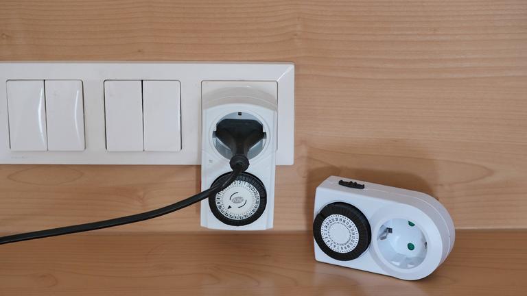 Eine Zeitschaltuhr für Steckdosen steckt an einer Leiste aus Steckdose und Lichtschaltern.