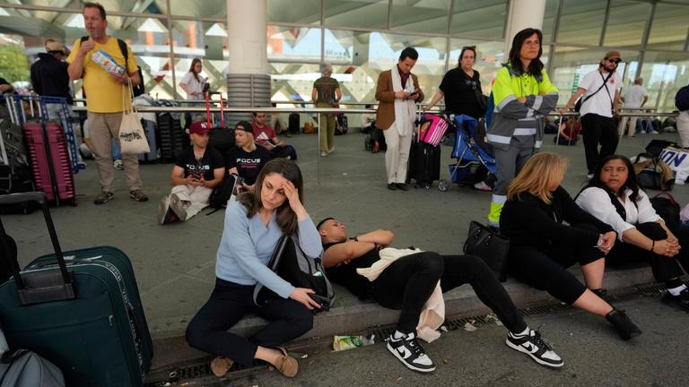 Menschen sitzen und stehen auf einem Bordstein vor dem Bahnhof Madrid-Atocha und warten.