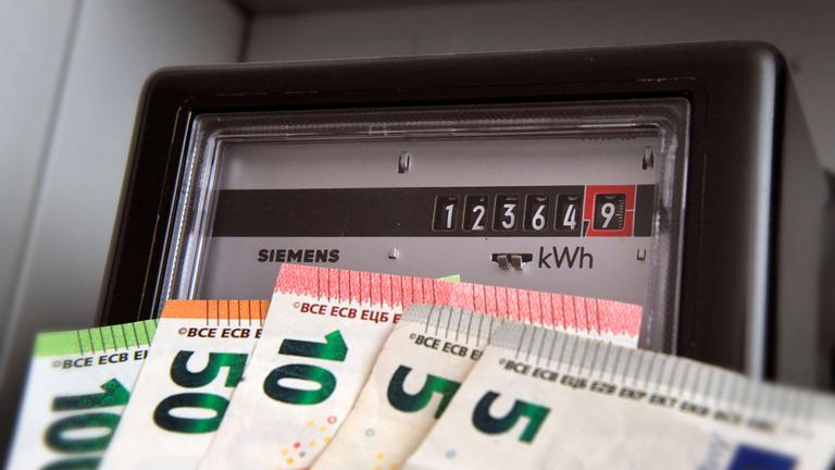 Symbolfoto: Ein Stromzähler mit Kilowattstundenzähler und verschiedenen Geldscheinen davor