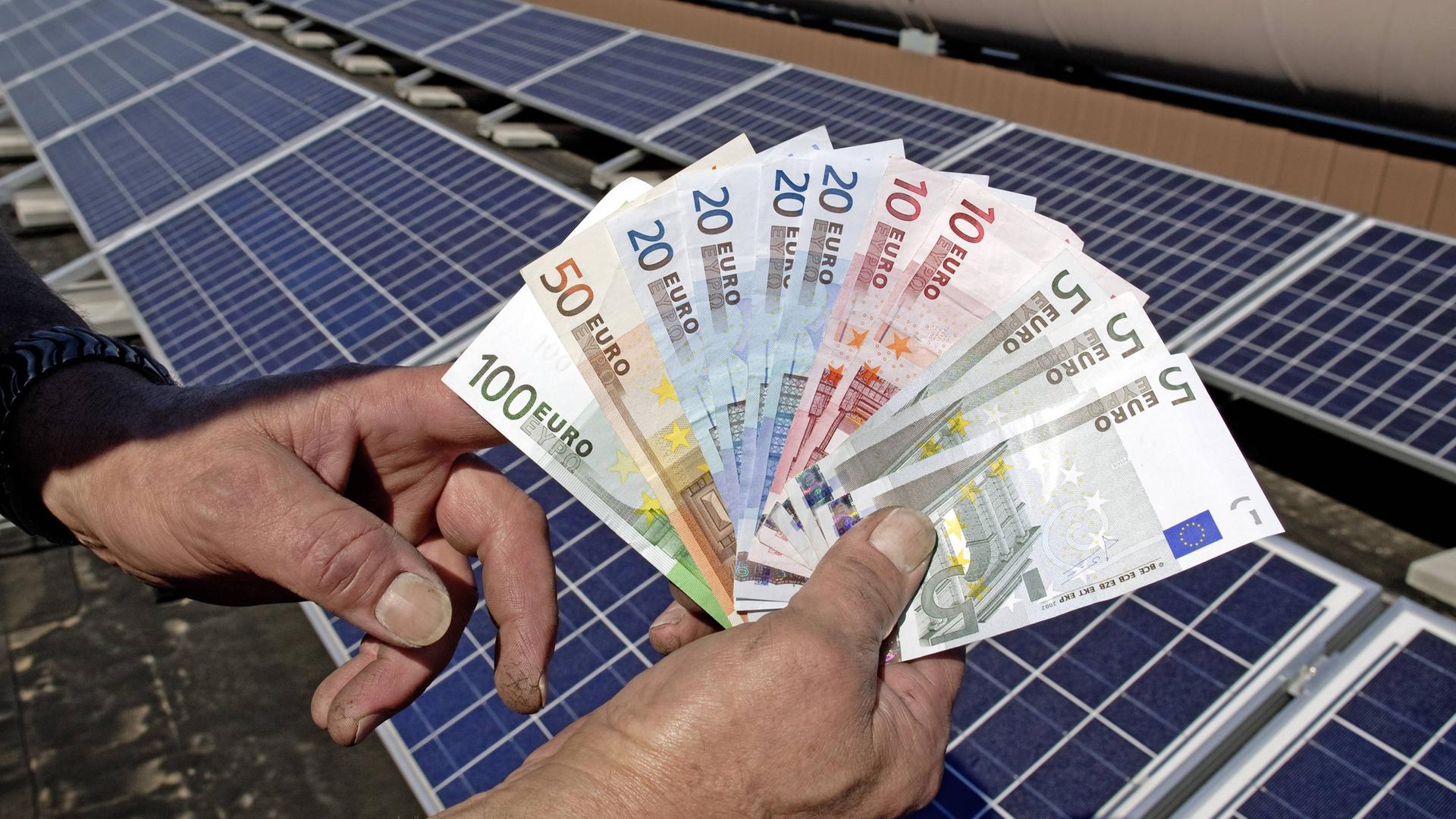 Eine Hand fächert Geldscheine auf - im Hintergrund sieht man Solarzellen (Symbolbild für Solarstrom).