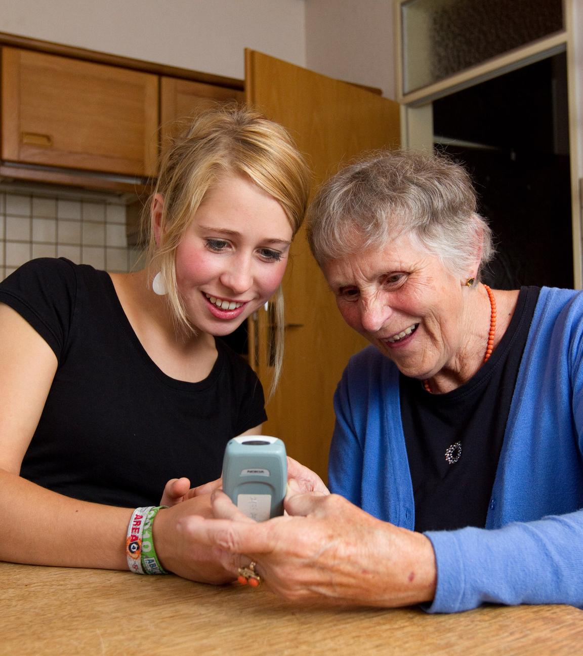 Eine Studentin sitzt mit einer Seniorin am Küchentisch und erklärt ihr die Benutzung des Handys.