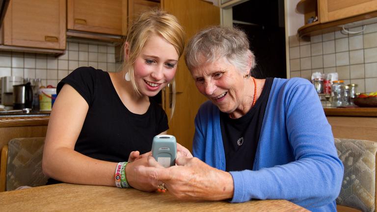 Eine Studentin sitzt mit einer Seniorin am Küchentisch und erklärt ihr die Benutzung des Handys.