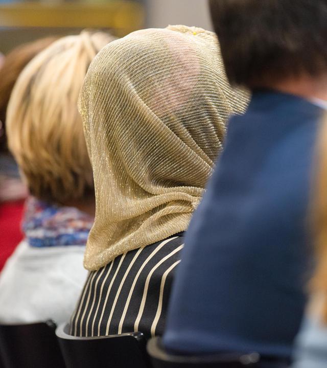 Mehrere Personen von hinten, sitzend in einer Reihe, unter ihnen eine Kopftuchträgerin