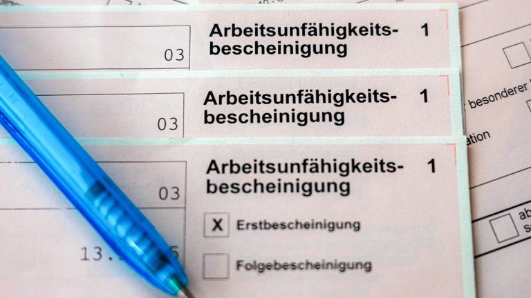 ILLUSTRATION - 18.02.2026, Bremen: Mehrere Exemplare einer Arbeitsunfähigkeitsbescheinigung liegen übereinander, daneben ein Kugelschreiber.