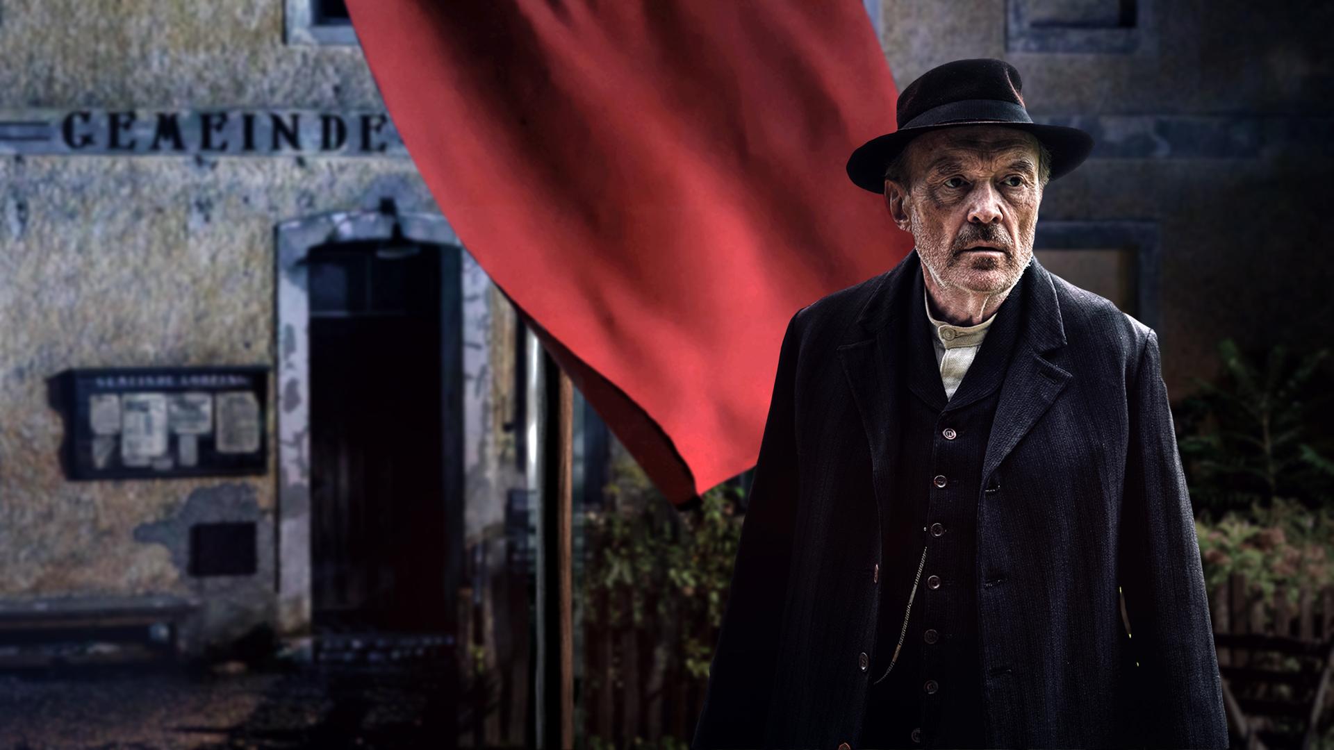 Einer der Hauptdarsteller steht nachts vor dem Rathaus einer Stadt. Hinter ihm weht eine große rote Fahne.