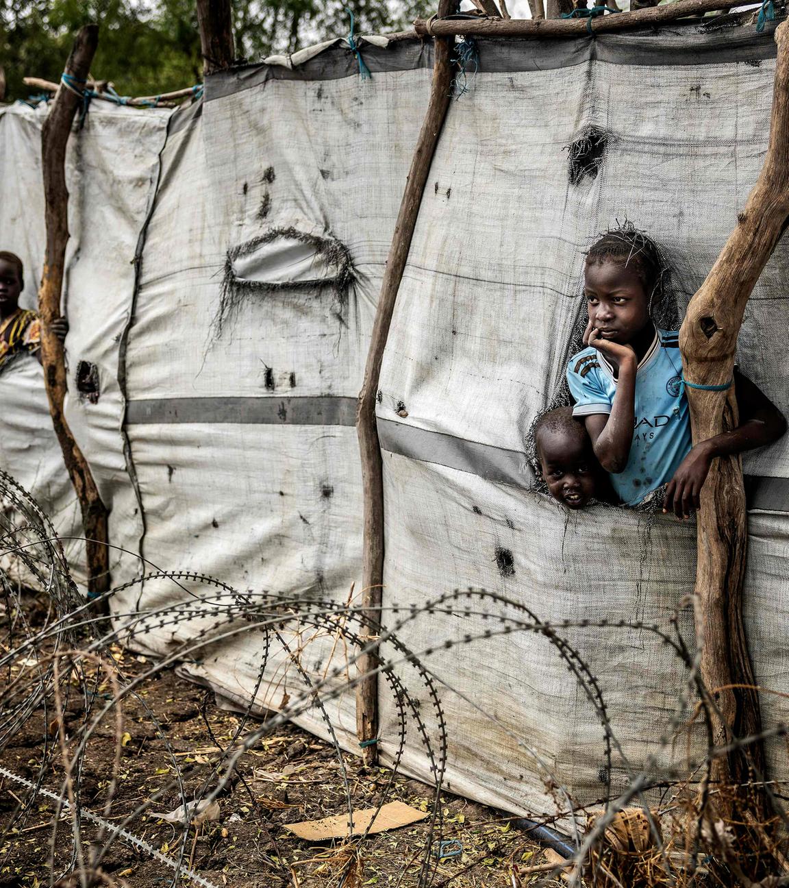 Kinder schauen aus Schlitzen in einer Plane am Zaun eines Flüchtlingslagers.