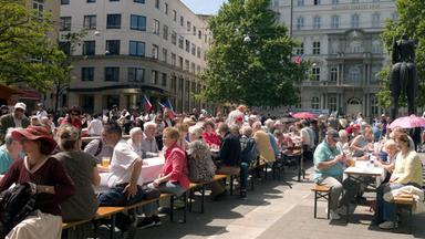 Auf einem Platz in Brünn sind lange Tischreihen aufgebaut. Die Leute sitzen auf Bierbänken davor.