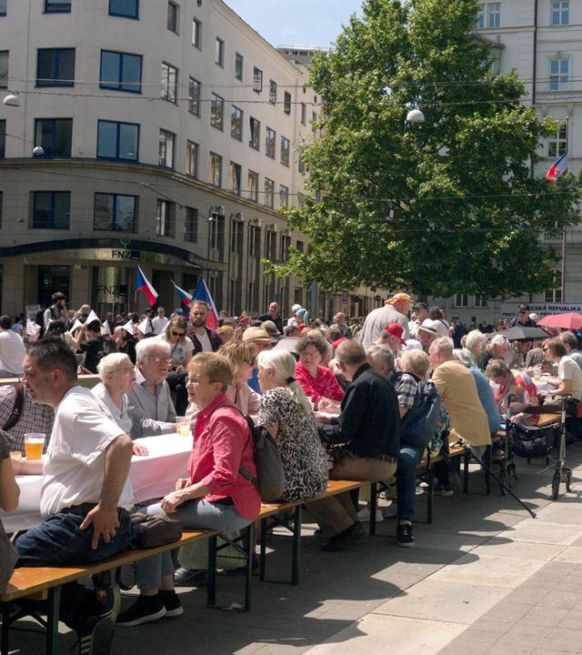 Auf einem Platz in Brünn sind lange Tischreihen aufgebaut. Die Leute sitzen auf Bierbänken davor.