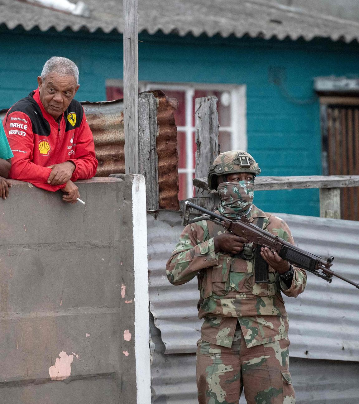 Ein Soldat mit Waffe in Tarnkleidung läuft durch eine Wohngegend. Dahinter lehnen zwei Personen an einem Zaun.