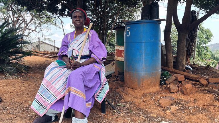 Eine afrikanische alte Frau sitzt draußen auf einem Hocker.