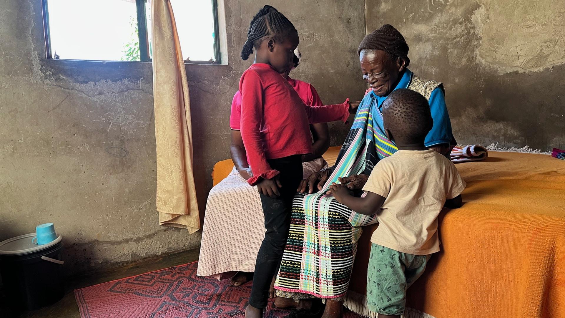 Eine alte Frau aus Südafrika sitzt auf einem Bett, neben ihr steht jeweils links und rechts ein Kind. Auf dem Bett sitzt noch eine Person, die verdeckt ist.