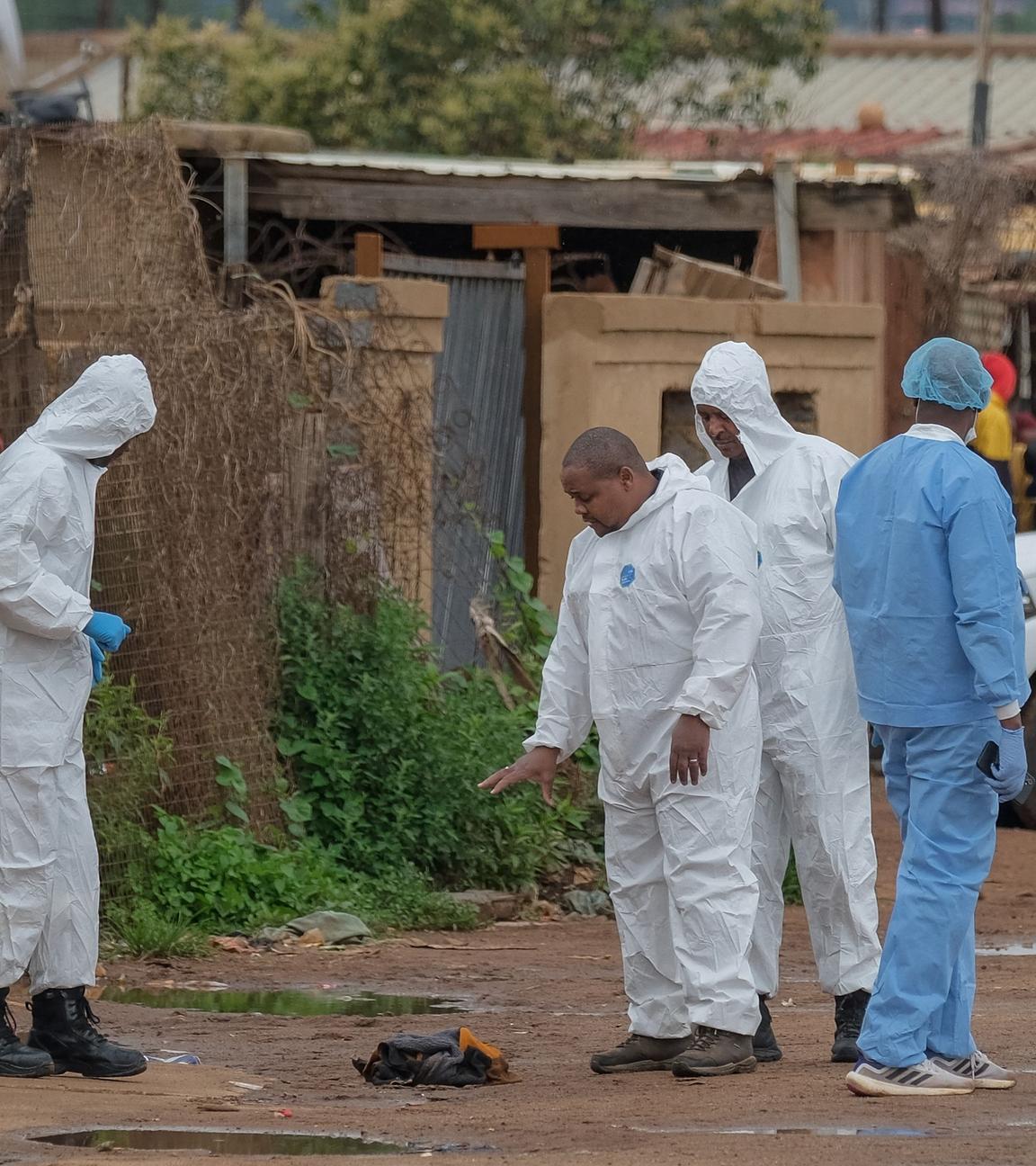 Mitglieder des Forensic Pathology Services der South African Police Service (SAPS) stehen am Tatort mit weißen Forensik-Anzügen, im Hintergrund ein weißer Jeep.