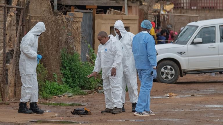 Mitglieder des Forensic Pathology Services der South African Police Service (SAPS) stehen am Tatort mit weißen Forensik-Anzügen, im Hintergrund ein weißer Jeep.