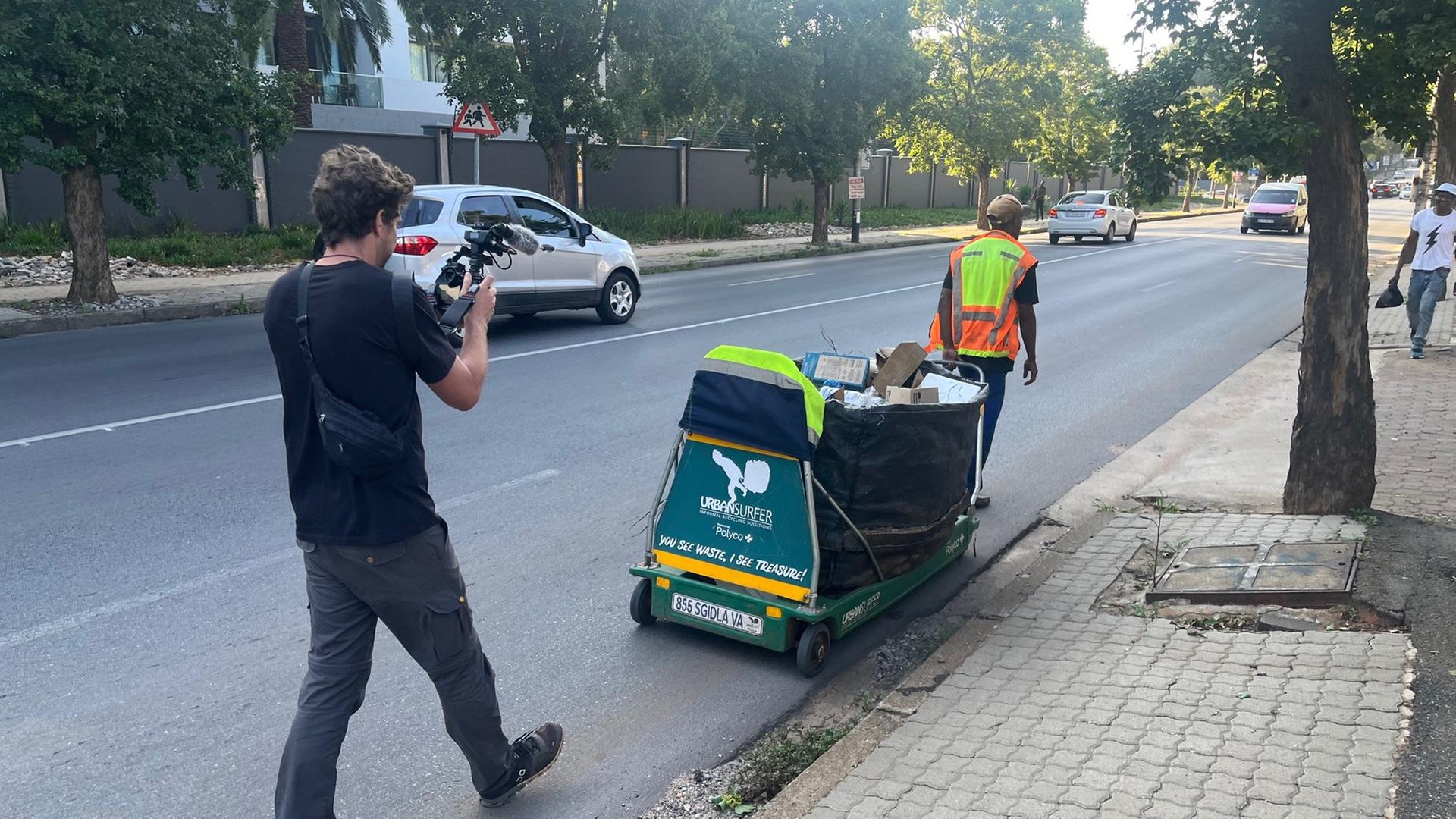 Auf dem Bild zu sehen ist ein Waste Picker mit Warnweste und einem Wagen für Müll. Hinter ihm ist ein Kameramann, der ihn begleitet.