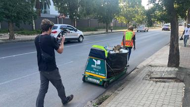 Auf dem Bild zu sehen ist ein Waste Picker mit Warnweste und einem Wagen für Müll. Hinter ihm ist ein Kameramann, der ihn begleitet.
