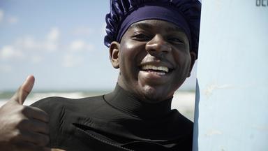 Mike Tsemo steht am Strand und schaut lachend in die Kamera, neben ihm ein Surfbrett.