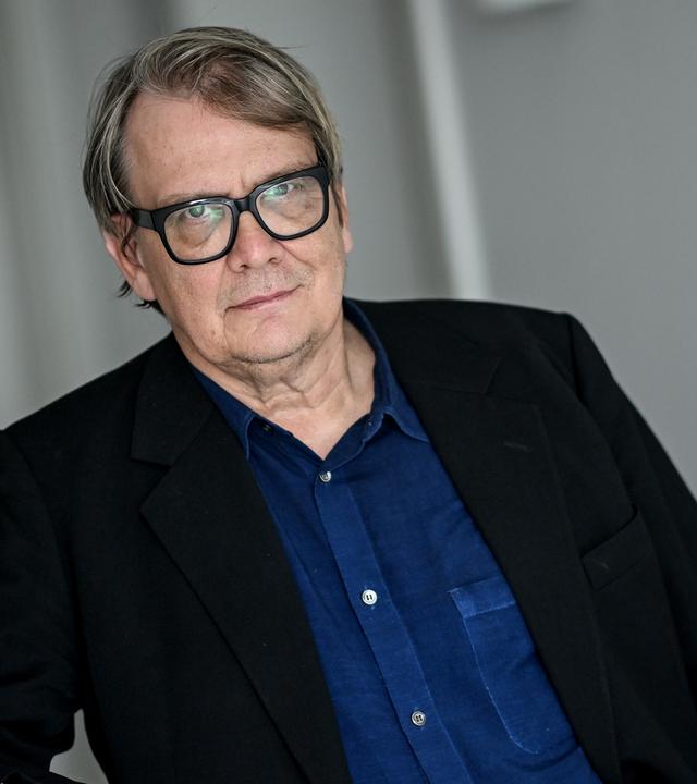 Porträtfoto eines Mannes im dunklen Anzug, der eine Brille mit dickem schwarzem Rahmen trägt.