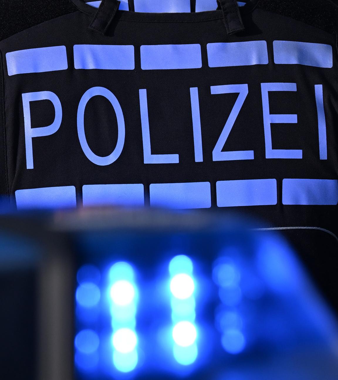 Das Logo der Polizei auf der Dienstkleidung eines Polizisten der vor einem Blaulicht steht. (Symbolfoto)