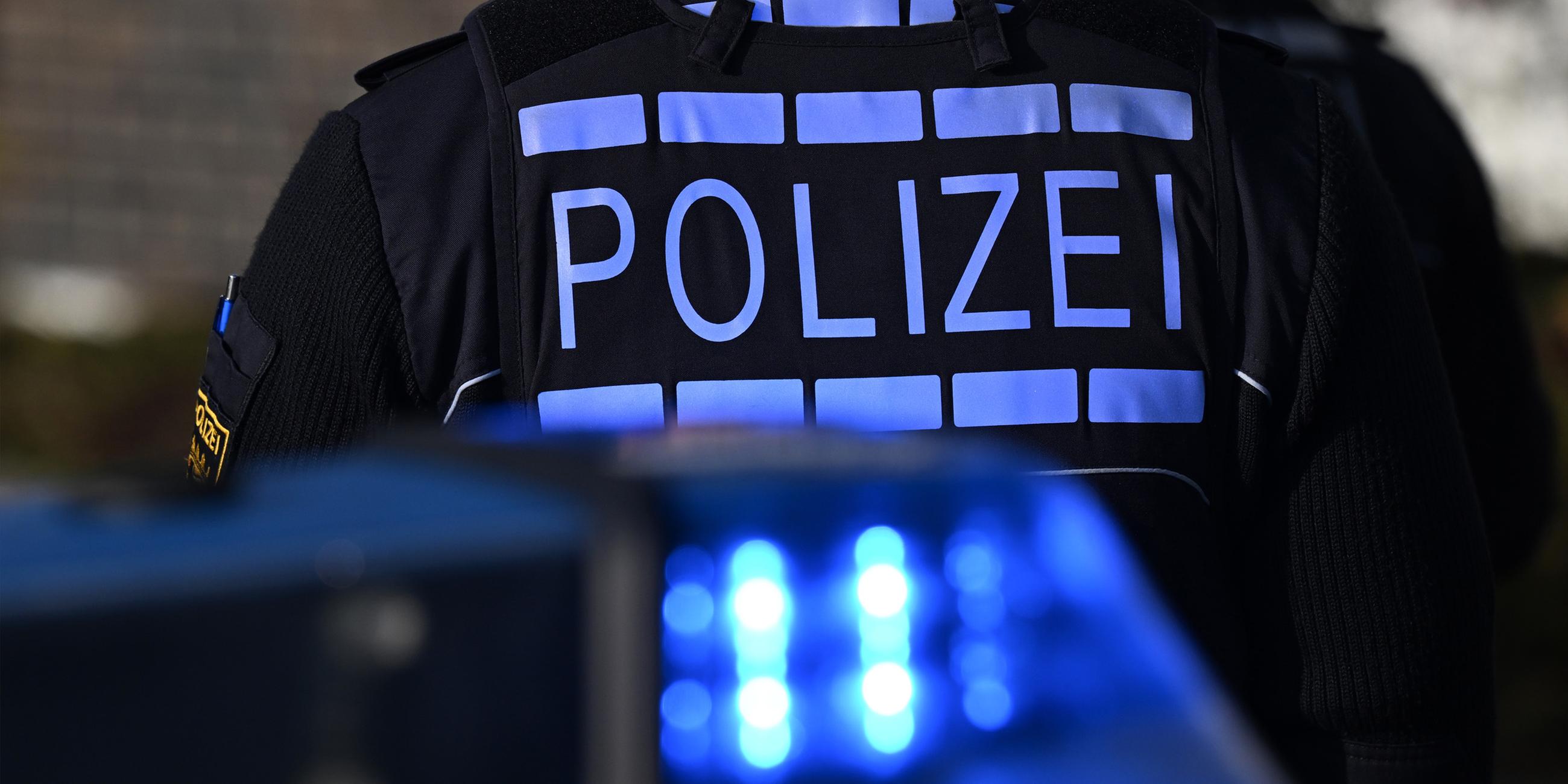 Das Logo der Polizei auf der Dienstkleidung eines Polizisten der vor einem Blaulicht steht. (Symbolfoto)