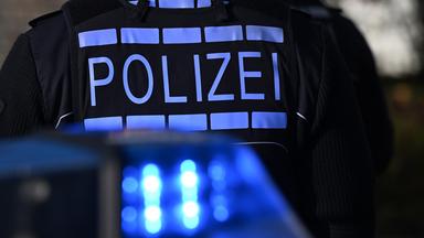 Das Logo der Polizei auf der Dienstkleidung eines Polizisten der vor einem Blaulicht steht. (Symbolfoto)