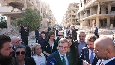 Johann Wadephul (M, CDU), Bundesminister des Auswärtigen, steht vor einer Straße mit während des Bürgerkriegs zerbombten Häusern.