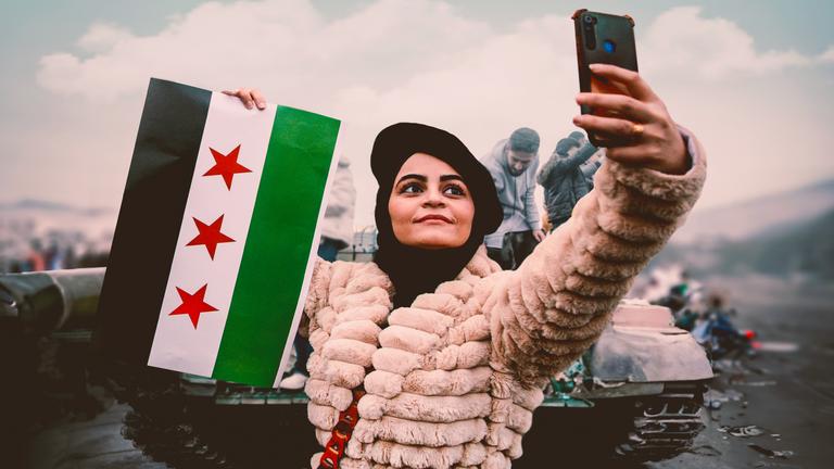Eine Frau macht ein Selfie. Sie hält in einer Hand ein Smartphone, in der anderen Hand eine syrische Fahne. Im Hintergrund befinden sich Männer auf einem Panzer.