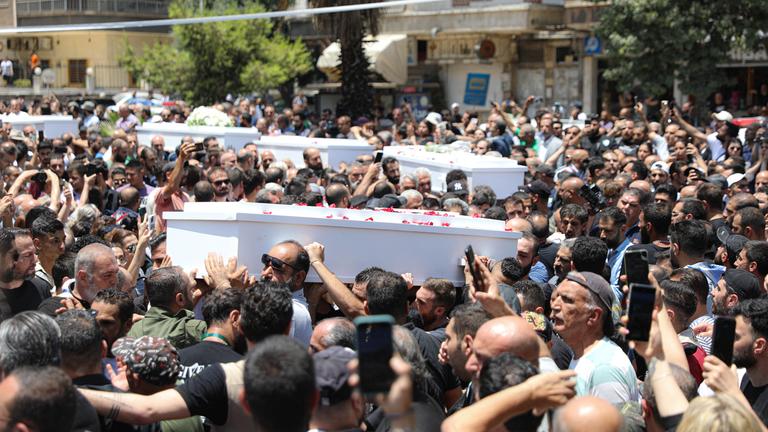 Weiße Särge werden durch eine Menschenmenge getragen, viele halten ihre Handys hoch, scheinbar, um das Ereignis zu dokumentieren.