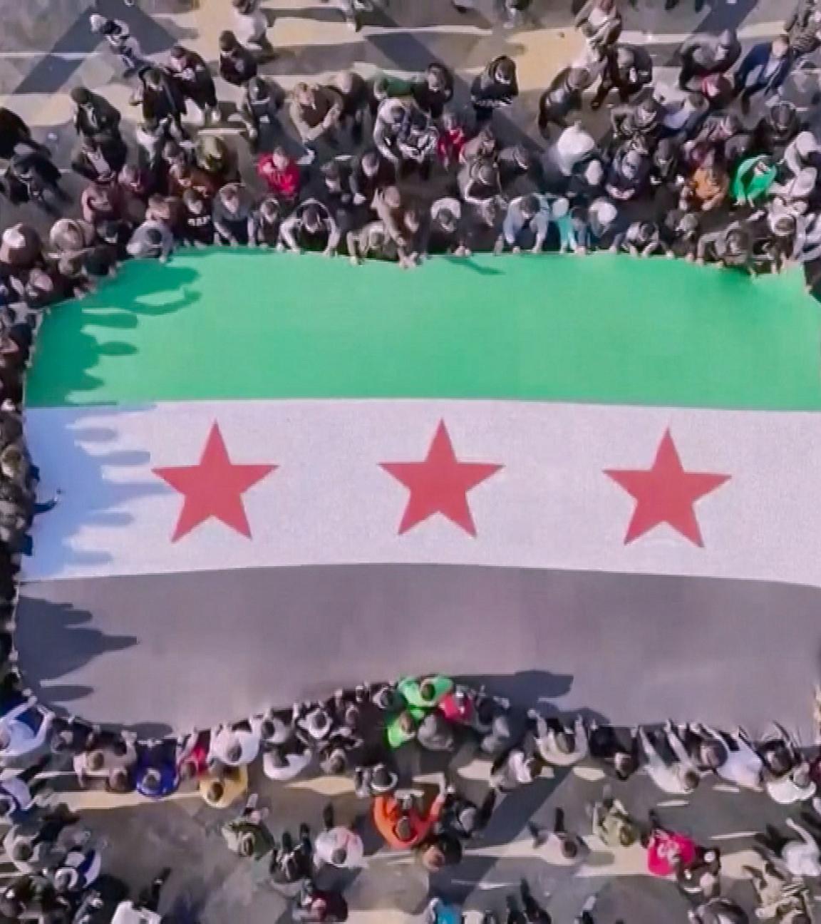 Vogelperspektive: Eine große syrische Fahne wird von einer Menschenmenge getragen. Die Fahne ist grün-weiß-schwarz gestreift mit drei roten Sternen in der Mitte.