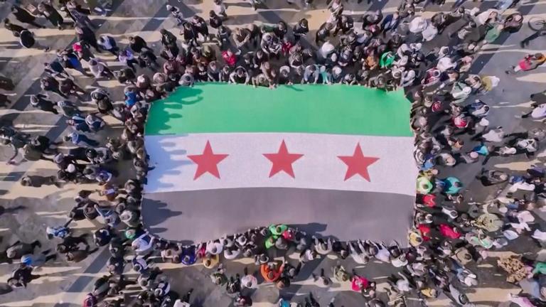 Vogelperspektive: Eine große syrische Fahne wird von einer Menschenmenge getragen. Die Fahne ist grün-weiß-schwarz gestreift mit drei roten Sternen in der Mitte.