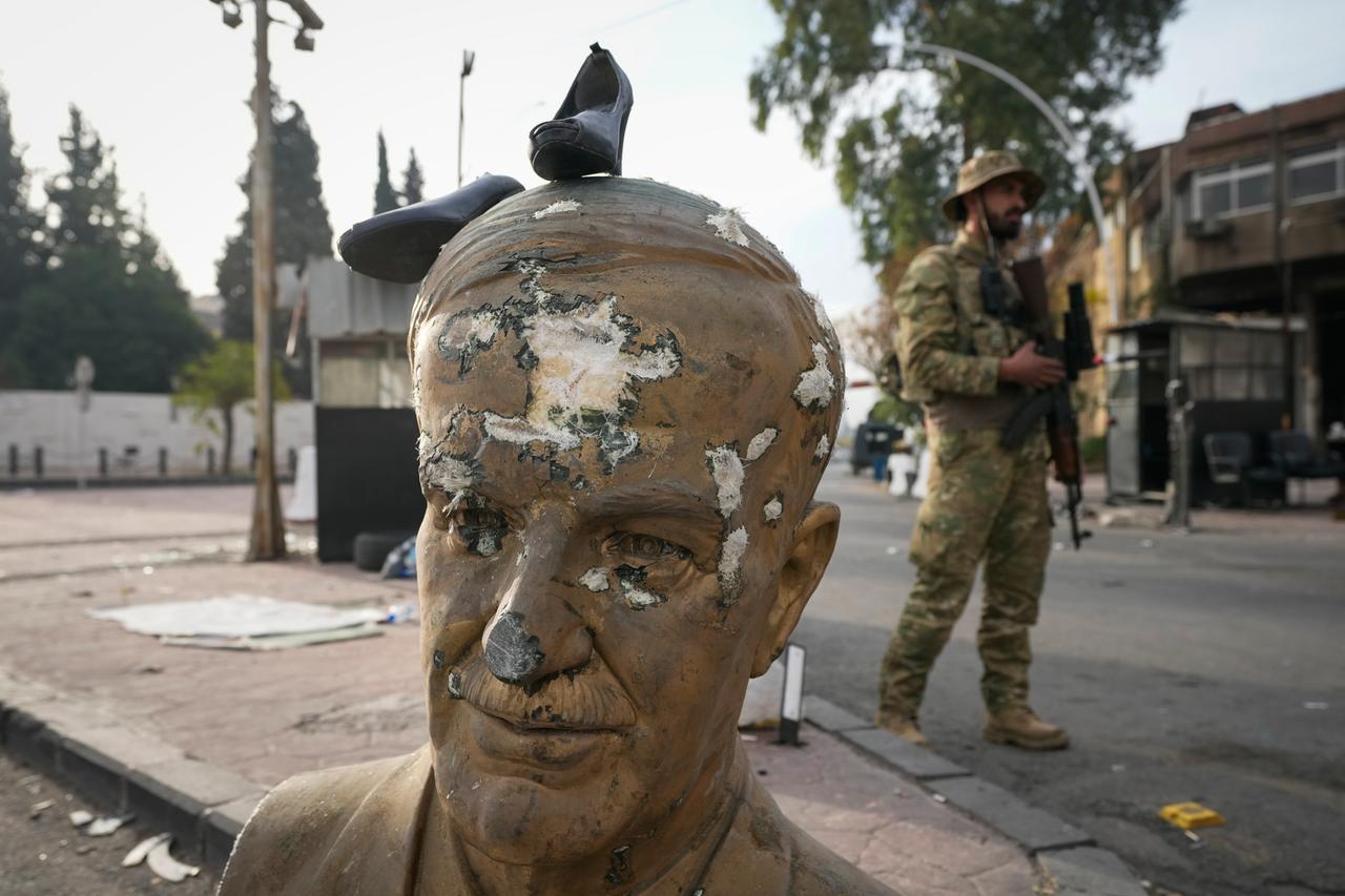 Ein Oppositionskämpfer in Militäruniform steht an einem Kontrollpunkt in Damaskus (Syrien) neben einer zerbrochenen Büste des verstorbenen syrischen Präsidenten Hafiz al-Assad, auf der ein Paar Schuhe liegt.