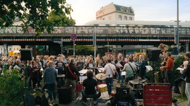 Im Vordergrund stehen eine Band und viele Menschen, die zusammen singen und Musik machen. Im Hintergrund ist eine Brücke zu sehen, über die ein Zug fährt.