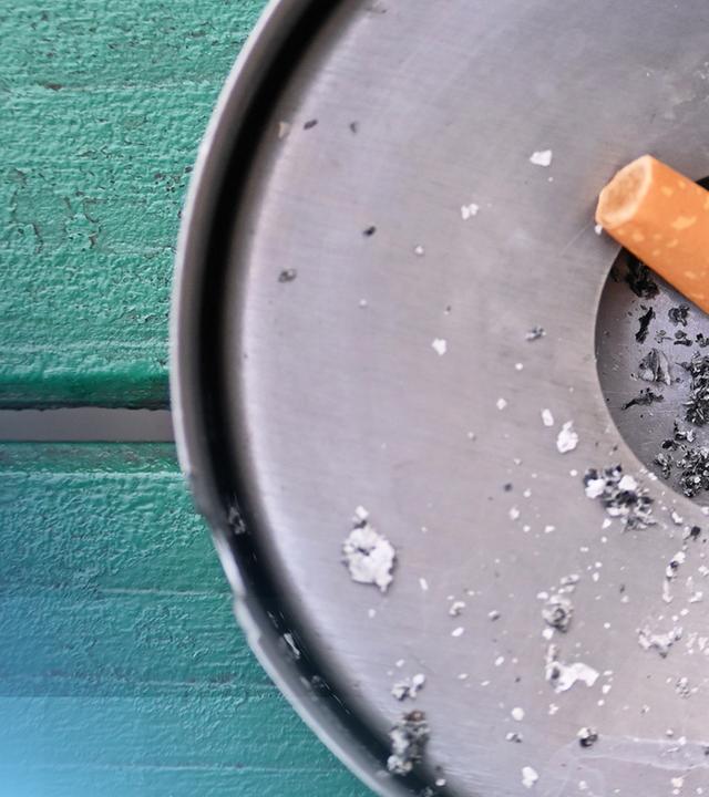 Kamerawinkel von oben: eine aufgerauchte Zigarette in einem metallenen Aschenbecher.