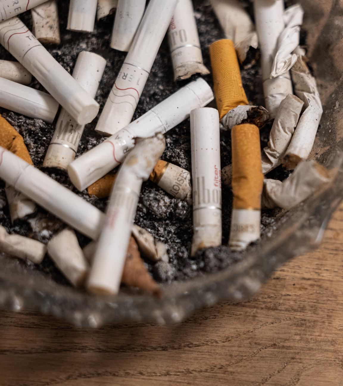 Ein Aschenbecher mit konsumierten Zigaretten und Tabaksticks steht auf einem Tisch. 