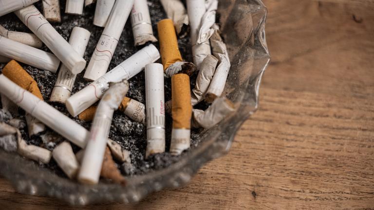 Ein Aschenbecher mit konsumierten Zigaretten und Tabaksticks steht auf einem Tisch. 