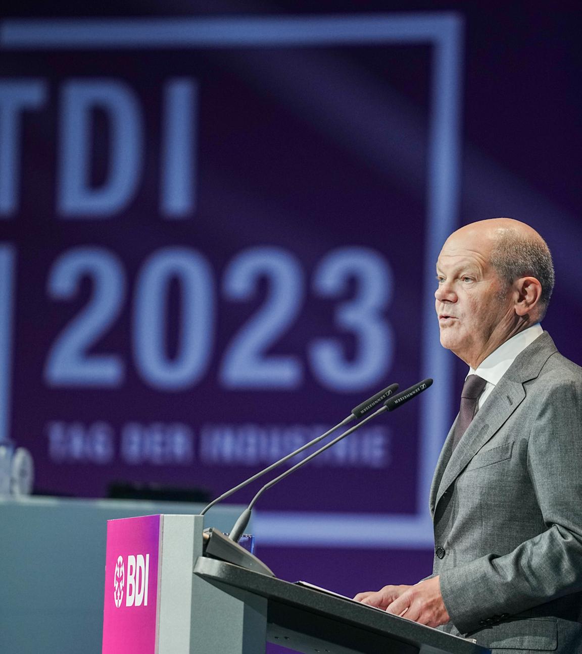 Der ehemalige Kanzler Olfa Scholz steht an einem Rednerpult. Im Hintergeund ist das Logo des Tages der deutschen Industrie zu sehen.