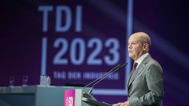 Der ehemalige Kanzler Olfa Scholz steht an einem Rednerpult. Im Hintergeund ist das Logo des Tages der deutschen Industrie zu sehen.