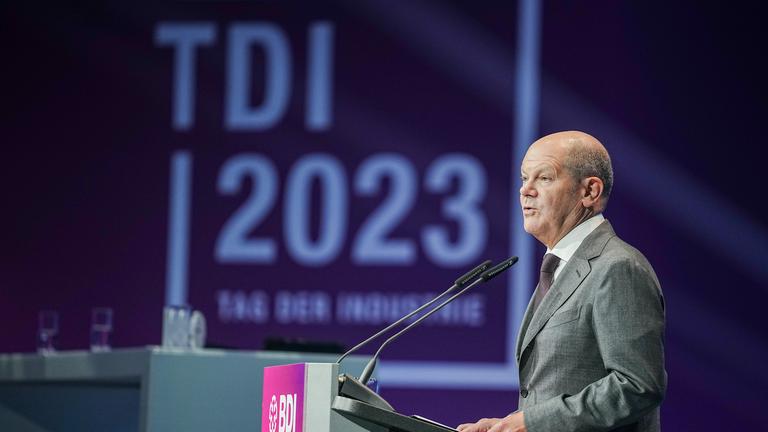Der ehemalige Kanzler Olfa Scholz steht an einem Rednerpult. Im Hintergeund ist das Logo des Tages der deutschen Industrie zu sehen.