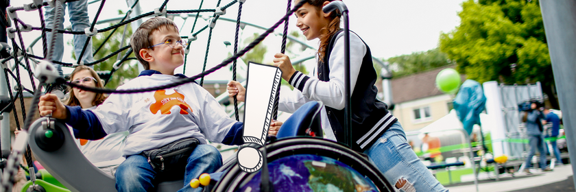 Ein Junge und ein Mädchen auf einem Spielplatz, davor steht ein Rollstuhl.