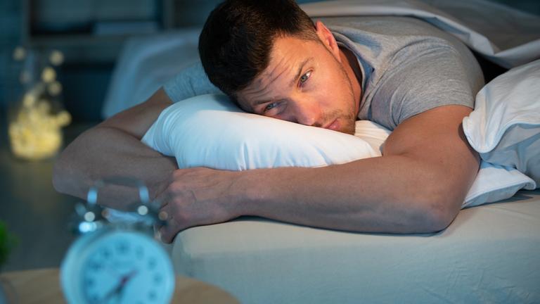 Ein junger Mann liegt auf dem Bauch im Bett und schaut geqäult seinen Wecker an.