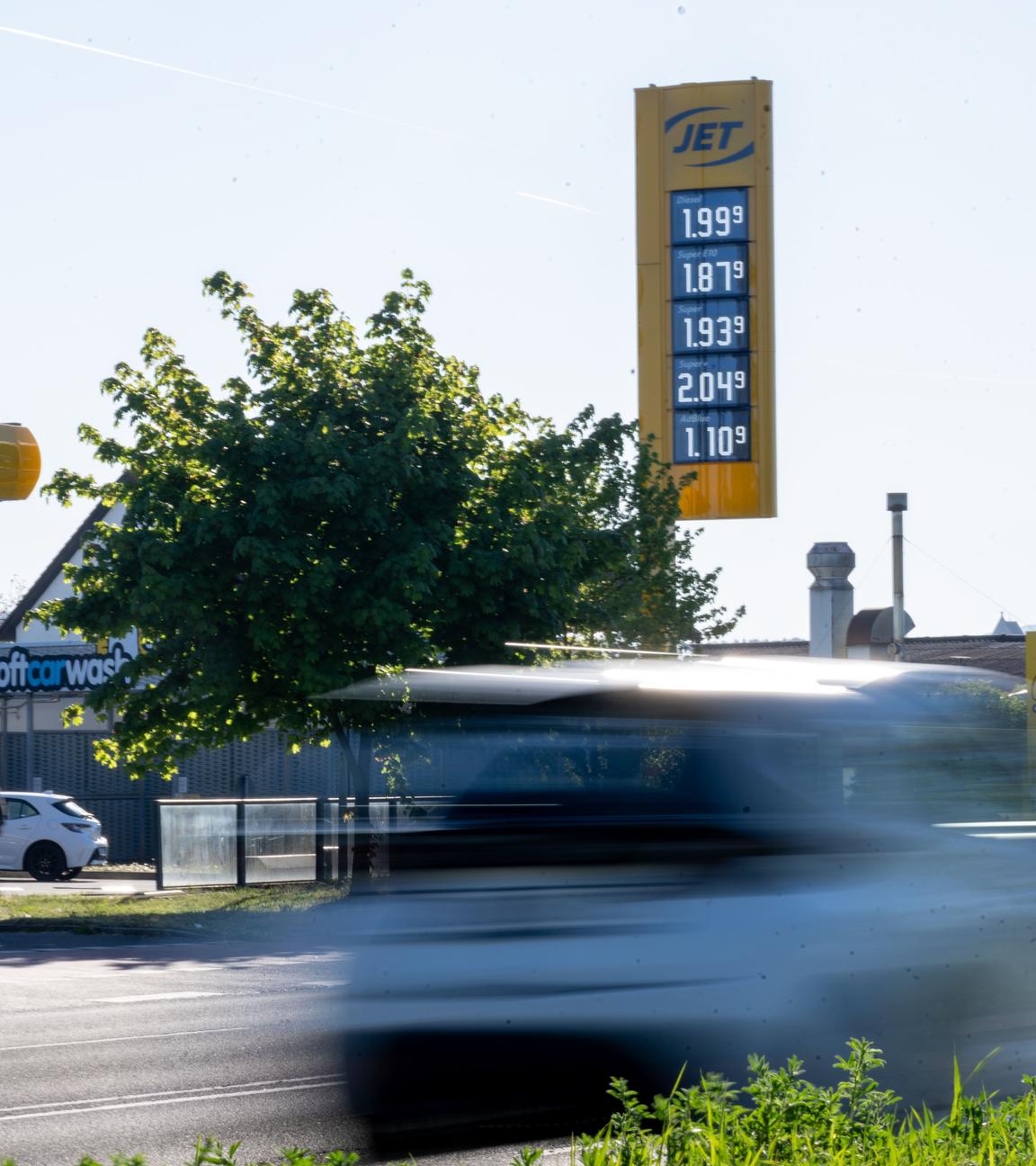 An einer Tankstelle in Bamberg stehen Autos zum Tanken an, während ein weiteres Fahrzeug auf einer Straße davor bei bestem Ausflugswetter vorbeifährt. Die Preisanzeige weist unter anderem dem Liter Diesel mit 1,999 Euro und den Liter Super E10 mit 1,879 Euro aus.