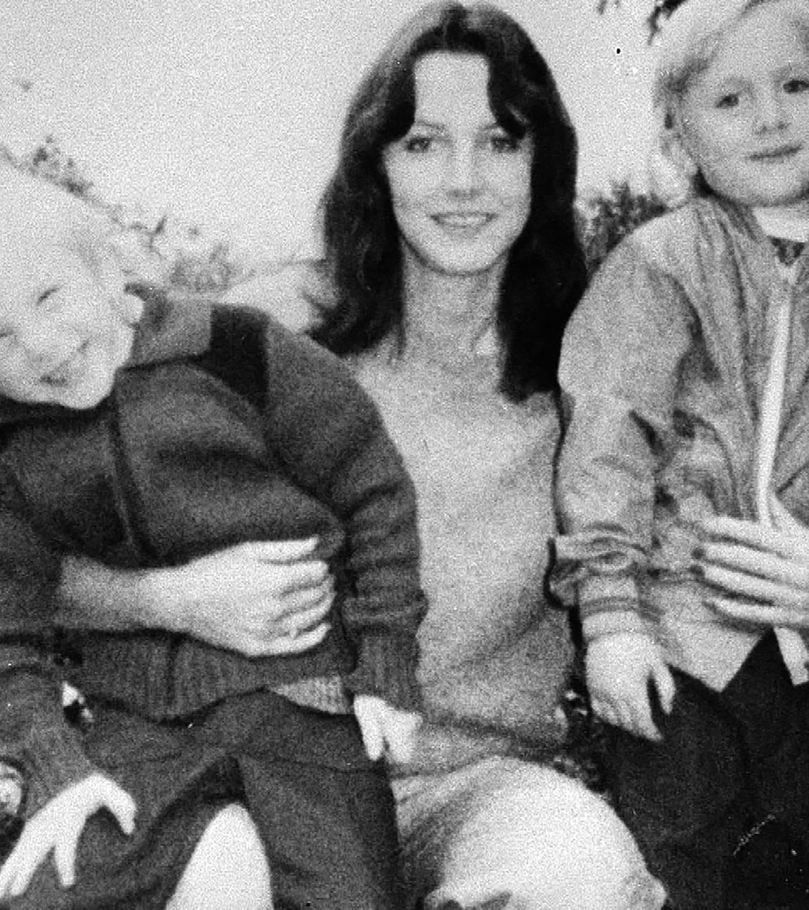 Schwarz-weiß-Foto einer jungen Frau. Sie hat lange braune, gewellte Haare und blickt mit einem leichten Lächeln in die Kamera. Auf ihrem Schoß sitzen zwei Jungen, ca. 5 Jahre alt. Die beiden sind Zwillinge.