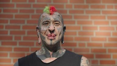 Tobias ist im ganzen Gesicht tätowiert und steht vor einer Ziegelsteinwand. An der Stirn hat er Hörner implantiert.