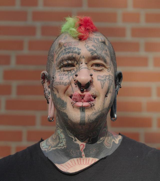Tobias ist im ganzen Gesicht tätowiert und steht vor einer Ziegelsteinwand. An der Stirn hat er Hörner implantiert.