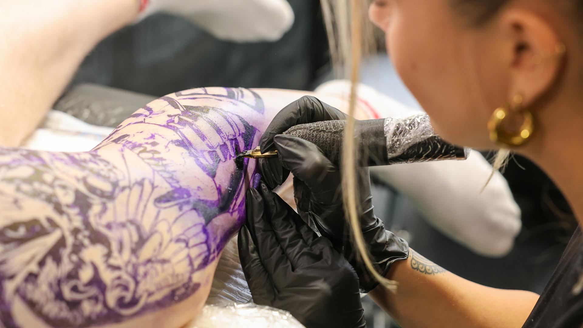 Eine Tätowiererin mit Einweghandschuhen und Tätowiergerät sticht ein großflächiges Bild auf einem Unterschenkel.