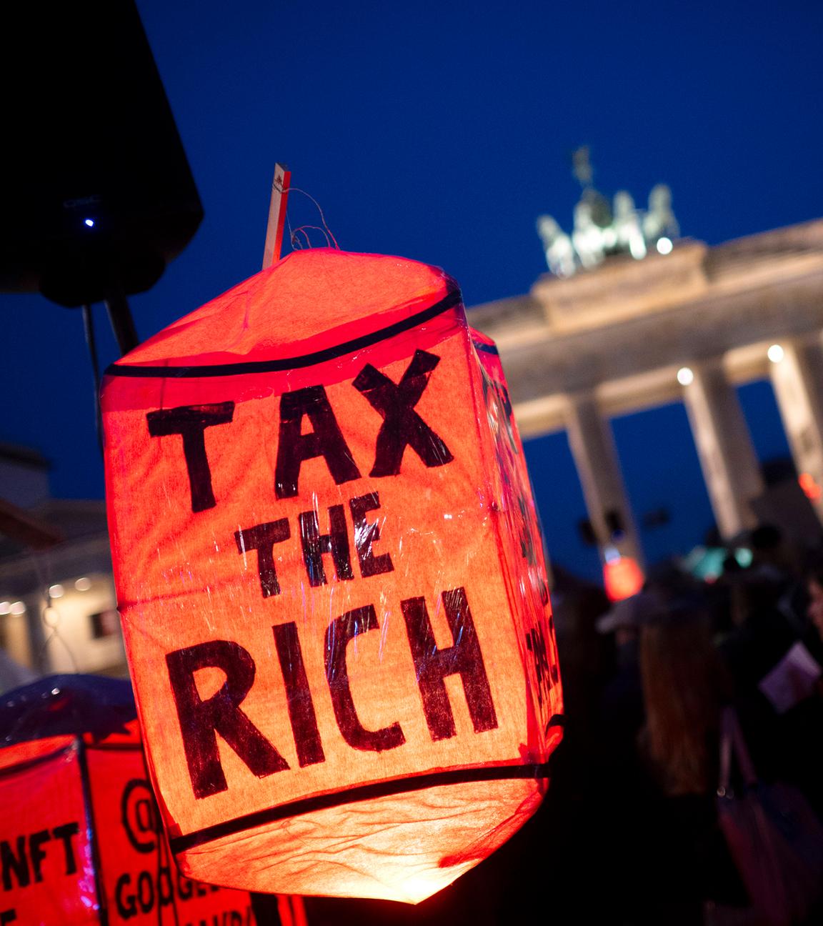 Bei einer Demo vor dem Brandenburger Tor bei Nacht ist im Vordergrund eine rote Lampe mit dem Text tax the rich zu sehen.