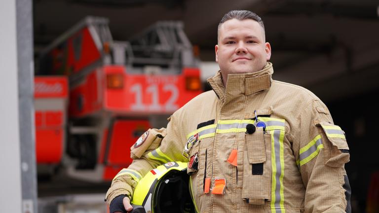 Junger mit mit Untercut in Feuerwehruniform steht vor einem Einsatzfahrzeug und lächelt in Richtung Kamera.