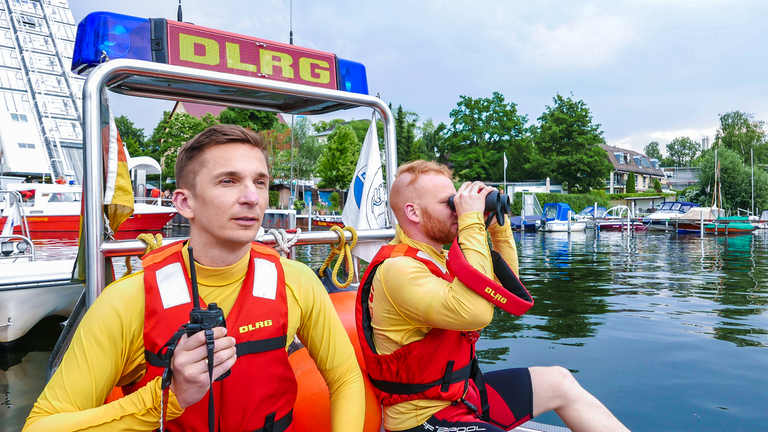 Eric sitzt zusammen mit einem Rettungschwimmer der DLRG in einem Boot der DLRG. Der Rettungsschwimmer schaut durchs Fernglas. Eric hät ein Fernglas in der Hand. Beide blicken auf den See hinaus.